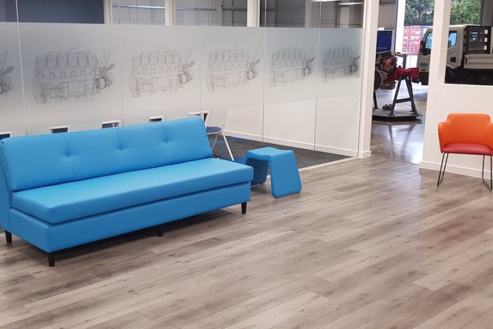 Office refurbishment with glazed screens with artwork, blue leather-effect couch and stool, orange chair and laminate flooring.
